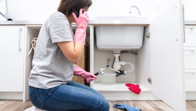 woman on the phone and holding a wrench to try and repair a kitchen sink leak